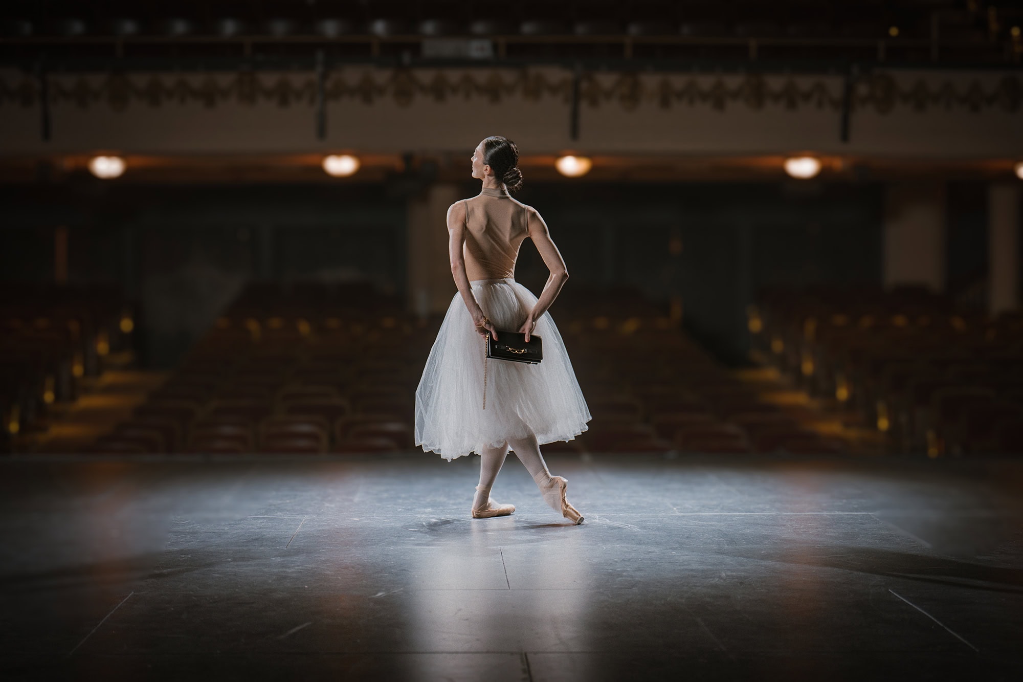 Ballet dancer in a white tutu holding a dark handbag on a dimly lit theater stage for AGCF Spring 2025 campaign.