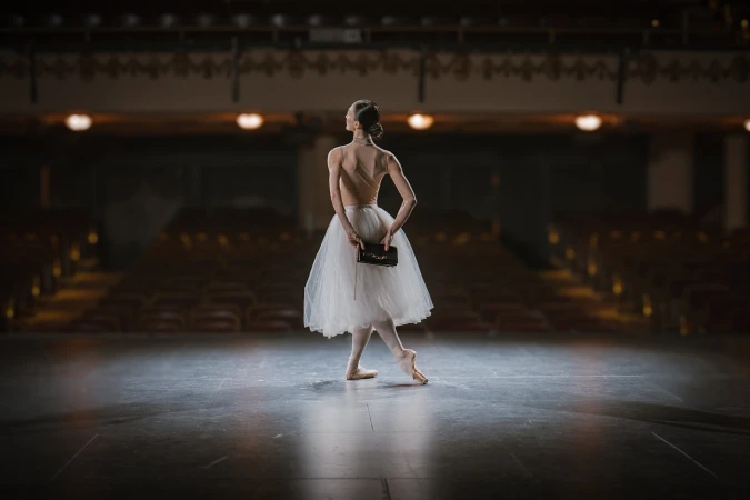 Ballet dancer in a white tutu holding a dark handbag on a dimly lit theater stage for AGCF Spring 2025 campaign.