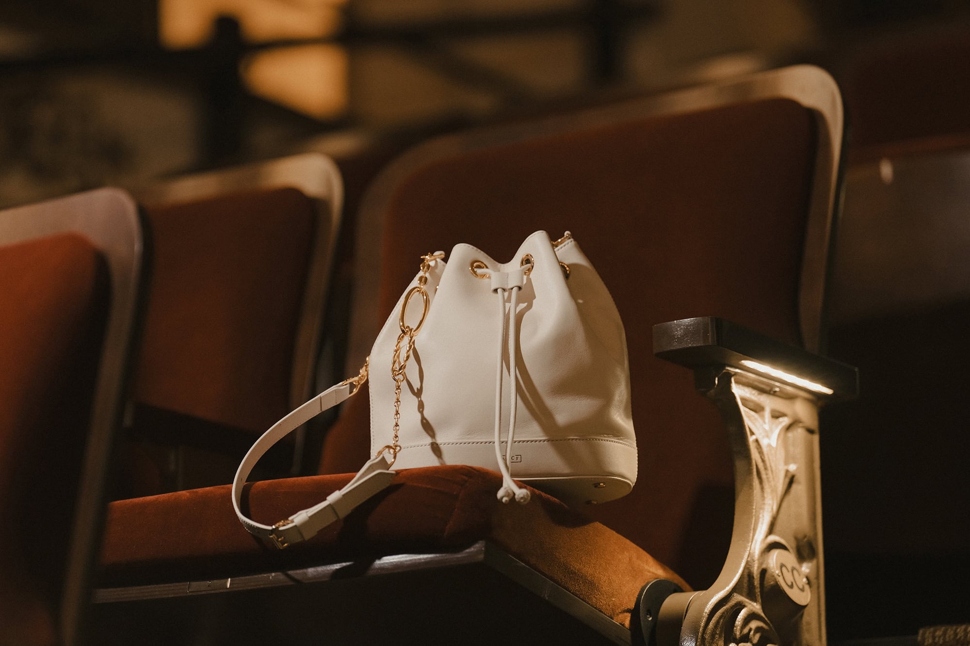 A white bucket bag rests on a plush red velvet theater seat within a dimly lit auditorium setting.