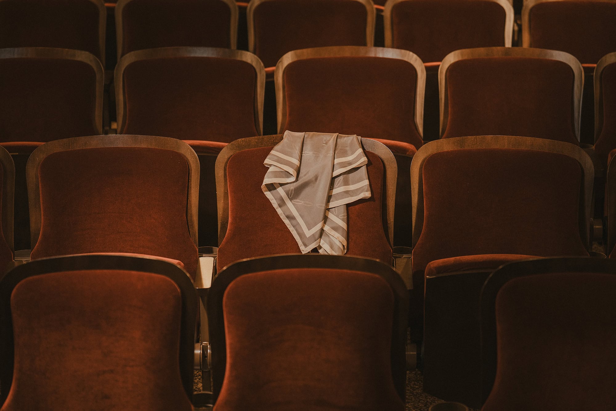 Folded grey and white striped silk scarf resting on a plush red velvet theatre seat among rows of empty auditorium chairs.