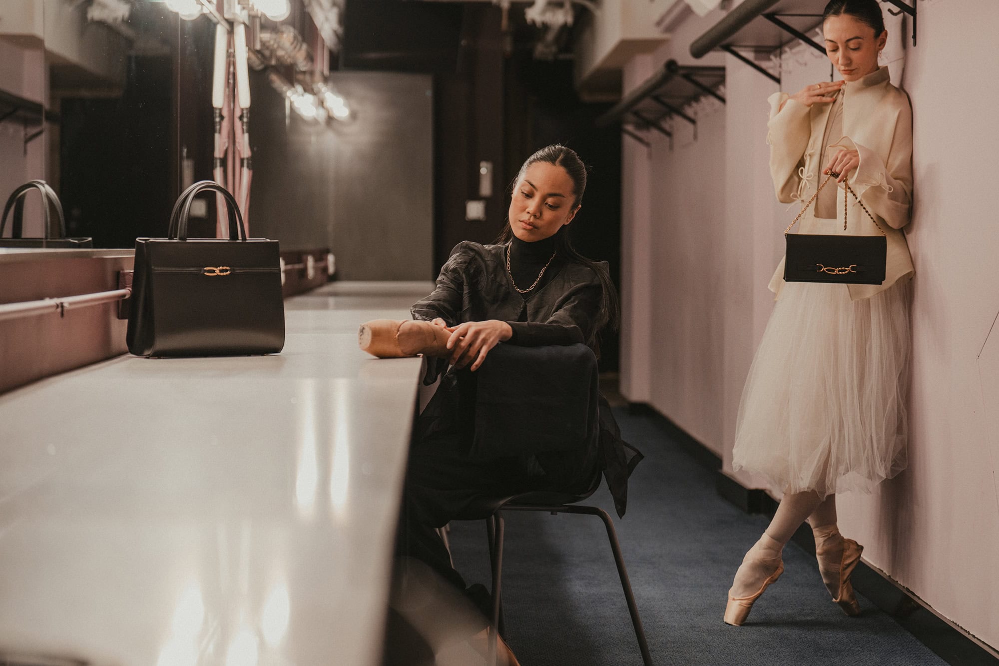 Two ballet dancers in a backstage dressing room with a black handbag resting on a vanity counter and another held by a dancer