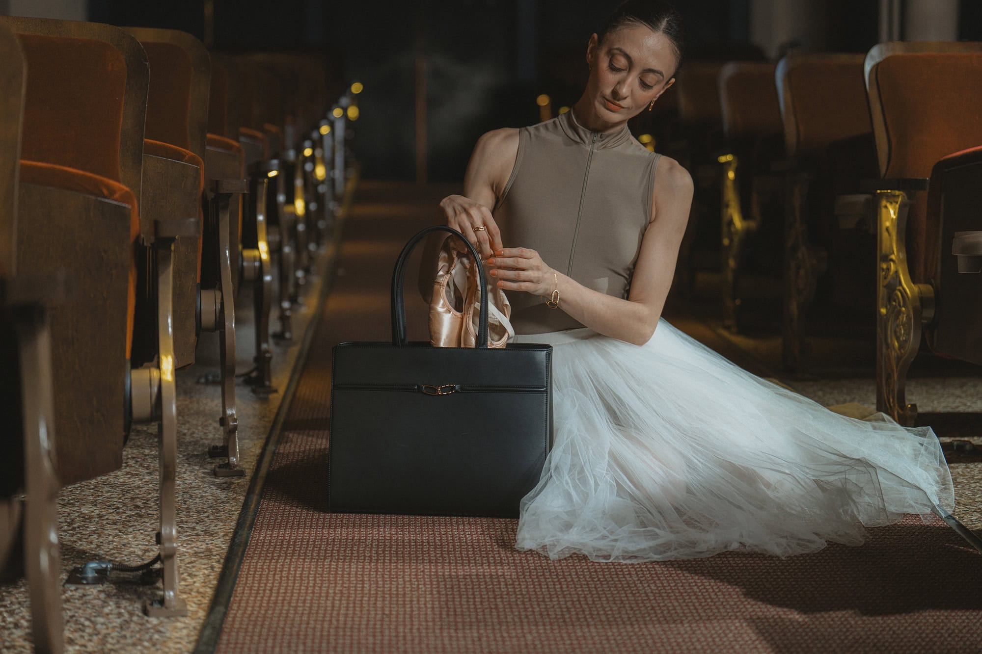 Dancer in a white tutu seated on a theater floor holding ballet shoes next to a black leather handbag.