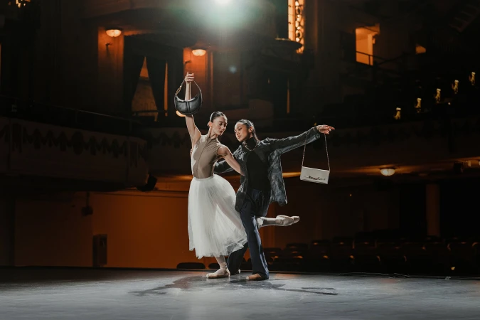 Two dancers posing on stage holding handbags inside a theatre interior with seating and warm lighting