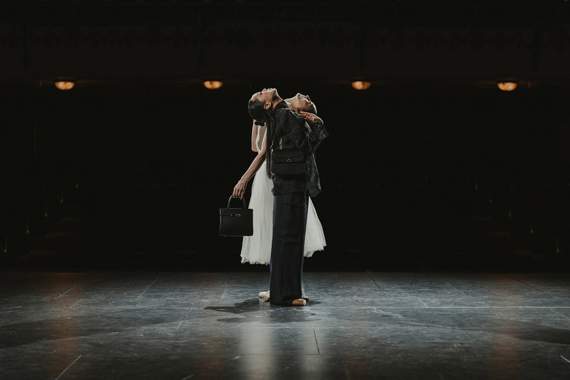 Two women dancing back to back on a dark stage holding black handbags for AGCF Spring 2025 campaign
