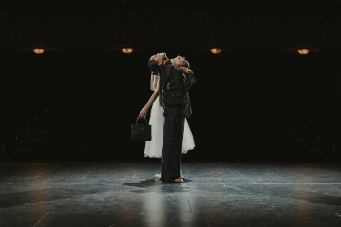 Two women dancing back to back on a dark stage holding black handbags for AGCF Spring 2025 campaign