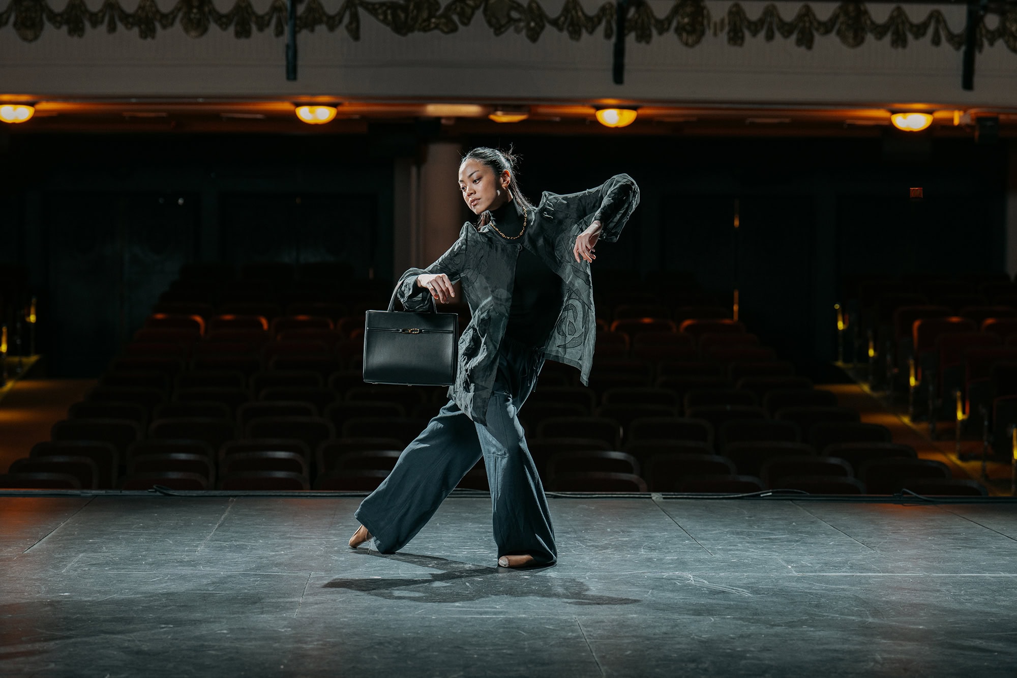 Dancer in a dark patterned jacket and wide trousers holding a black handbag on an empty theater stage.