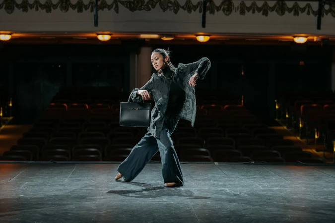 Dancer in a dark patterned jacket and wide trousers holding a black handbag on an empty theater stage.