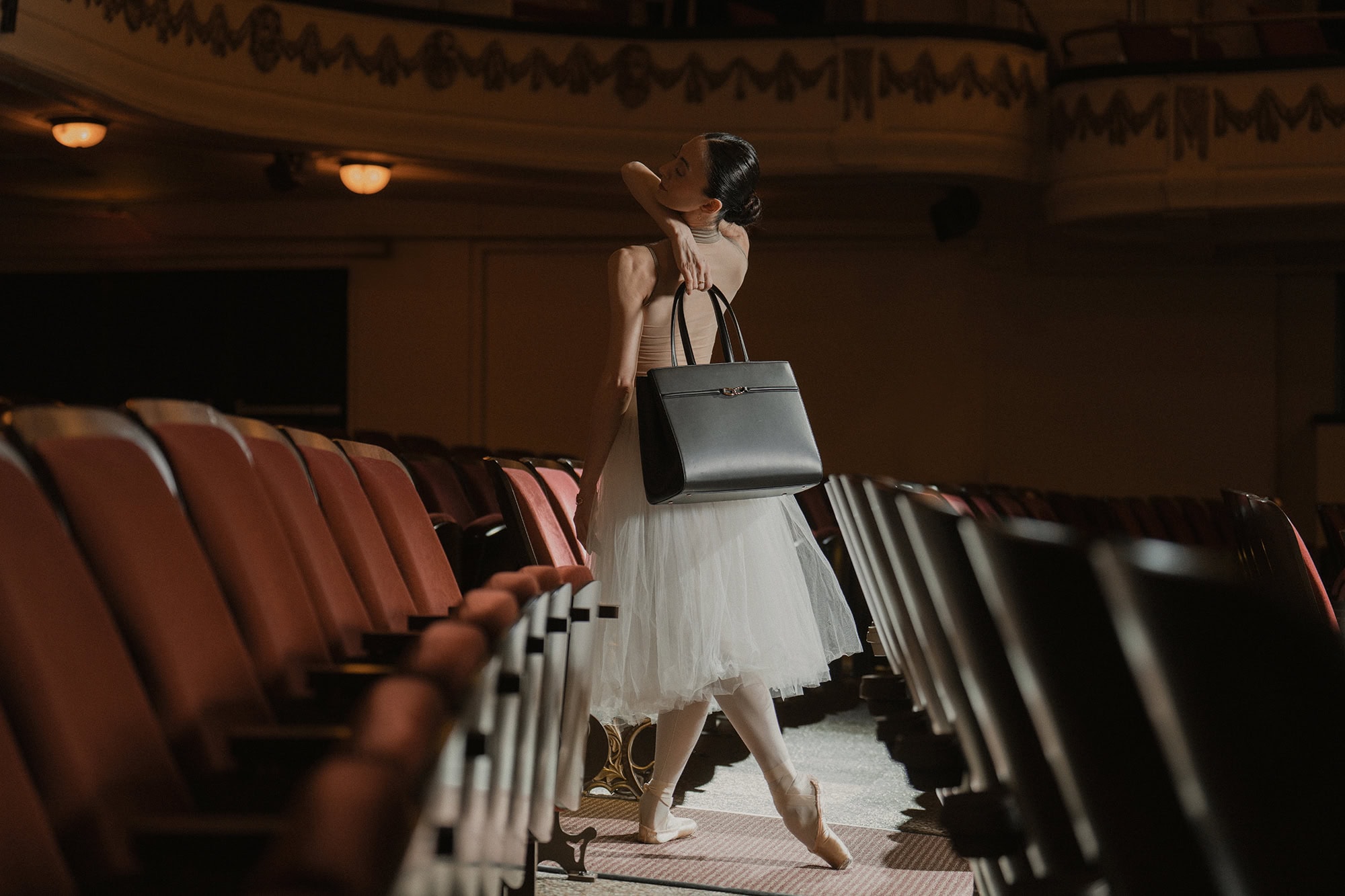 Ballet dancer wearing a white tutu holding a black handbag inside a historic theatre with rows of empty red seats.