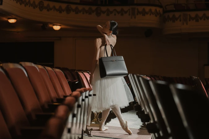 Ballet dancer wearing a white tutu holding a black handbag inside a historic theatre with rows of empty red seats.