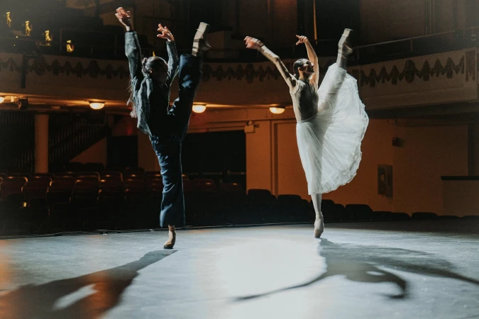 Two ballerinas dancing on stage in a theatre, one in dark clothing and one in a white skirt, with dramatic stage lighting.