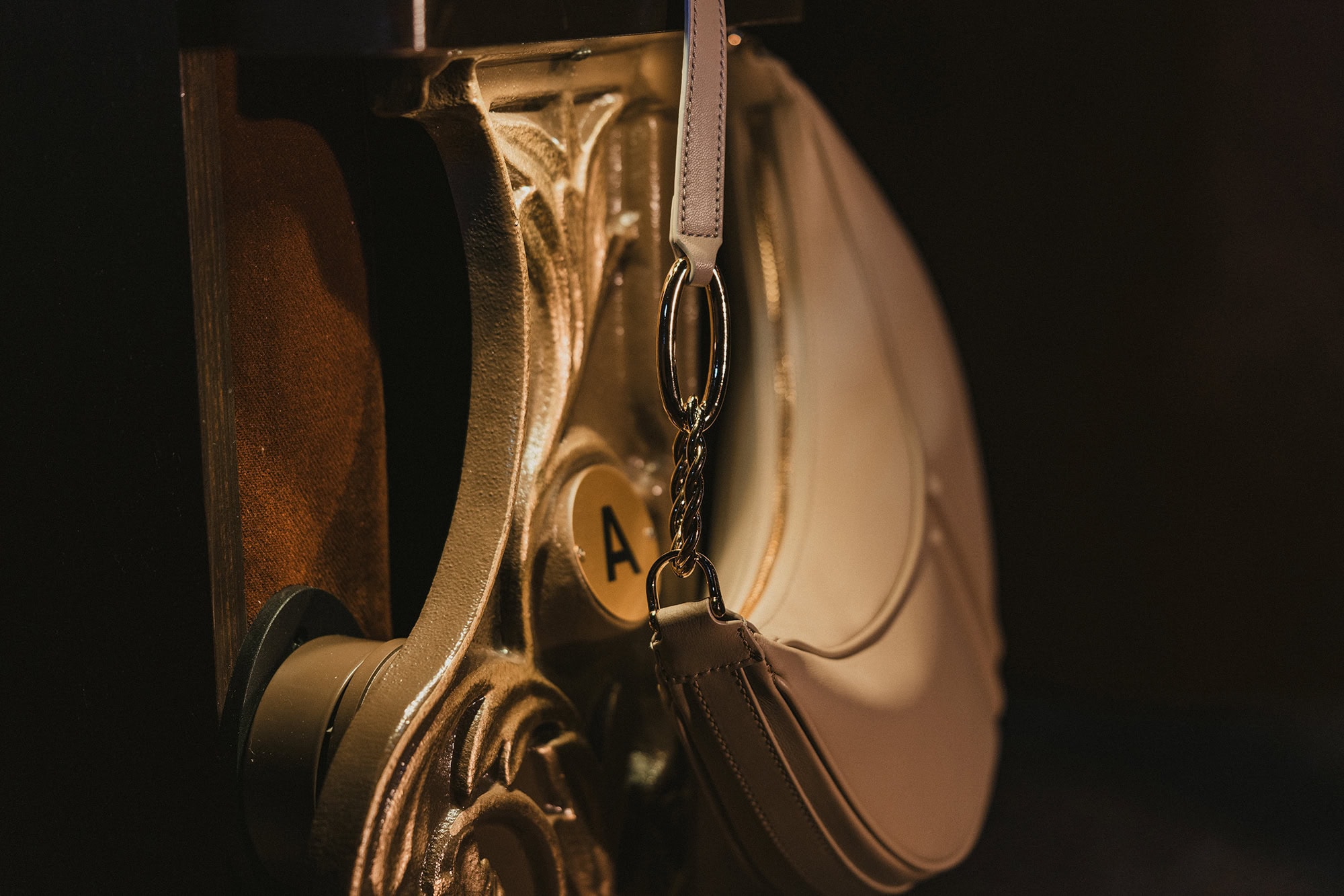 Beige leather shoulder bag hanging from an ornate gold architectural bracket for AGCF ad campaign.