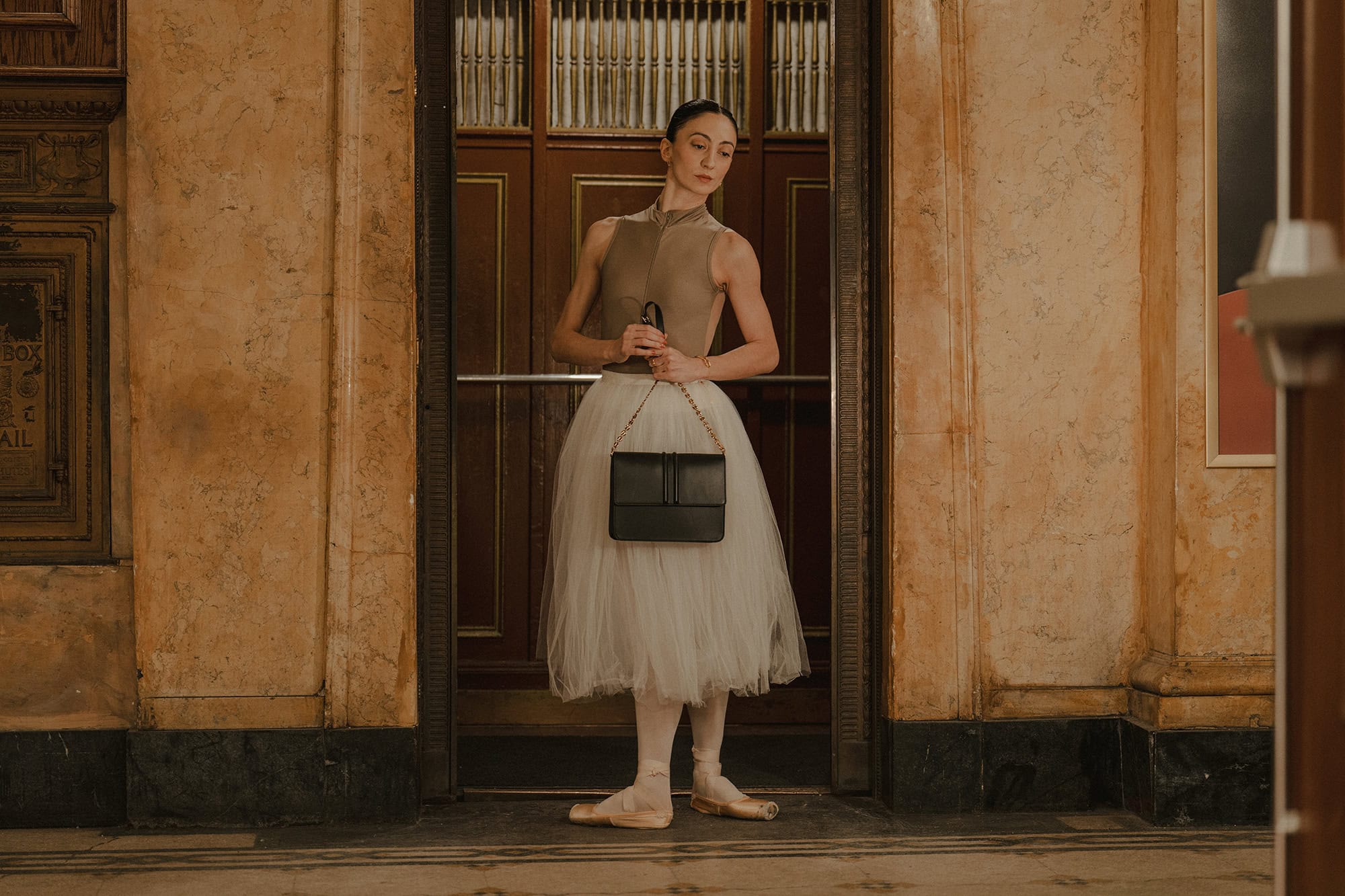 Dancer in a grey leotard and white tutu holding a black handbag standing inside a vintage elevator entrance.