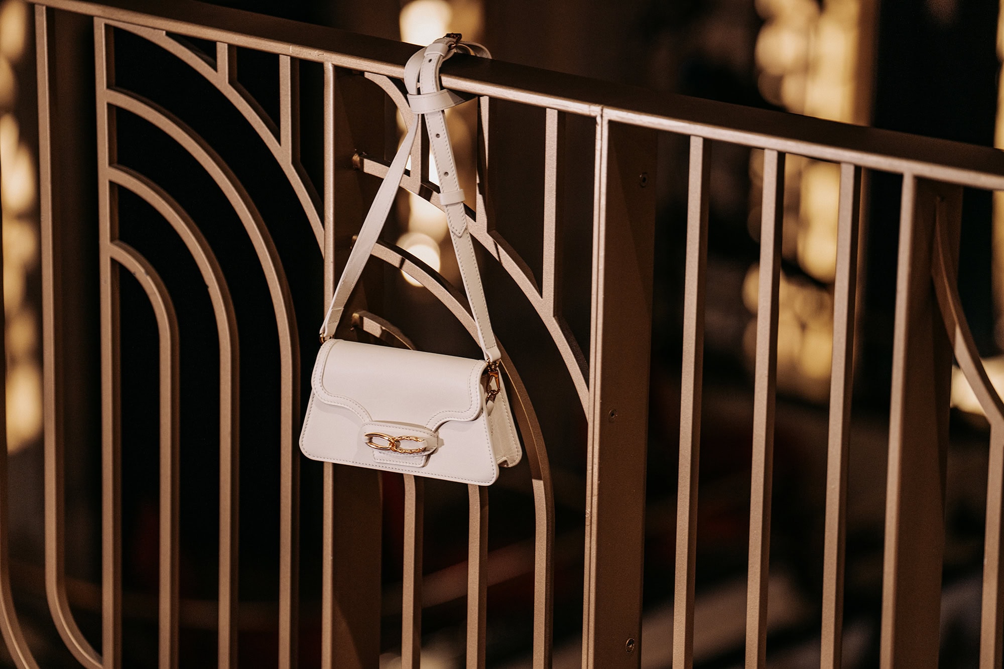 White leather handbag hanging from an ornate metal balcony railing with warm background lighting