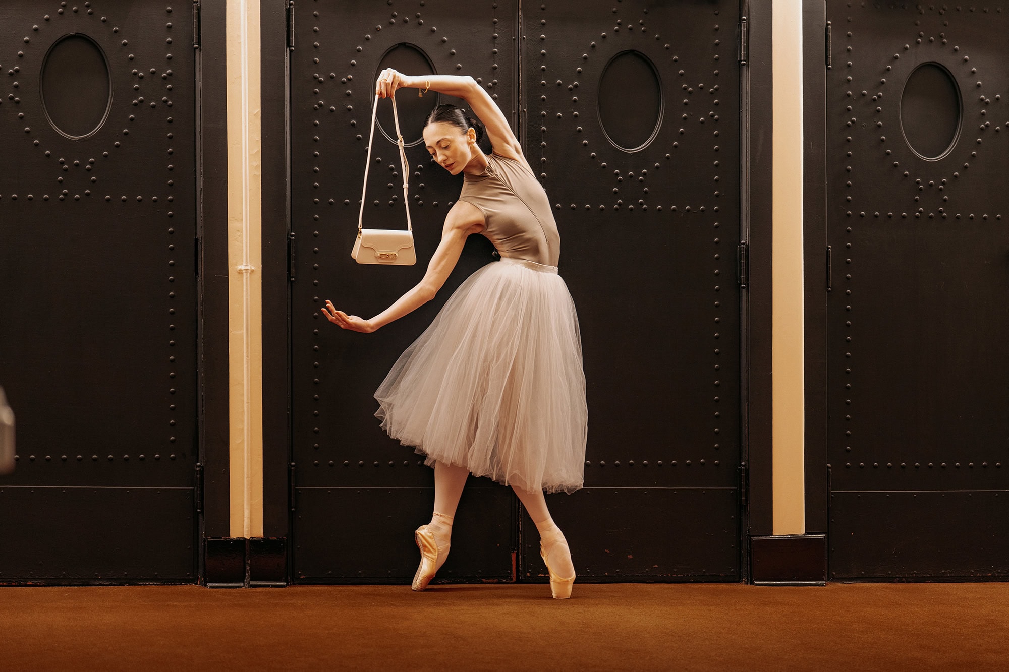Dancer in a tulle skirt posing en pointe holding a handbag in front of heavy dark double doors for AGCF Spring 2025 ad campaign.