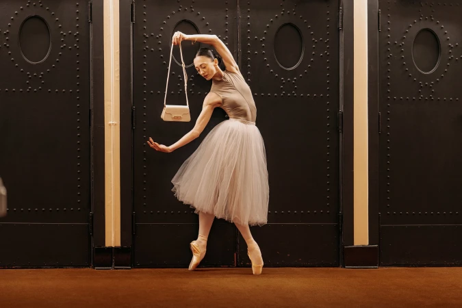 Dancer in a tulle skirt posing en pointe holding a handbag in front of heavy dark double doors for AGCF Spring 2025 ad campaign.