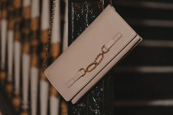 Beige leather shoulder bag with gold chain strap and interlocking metal hardware against a dark architectural background.