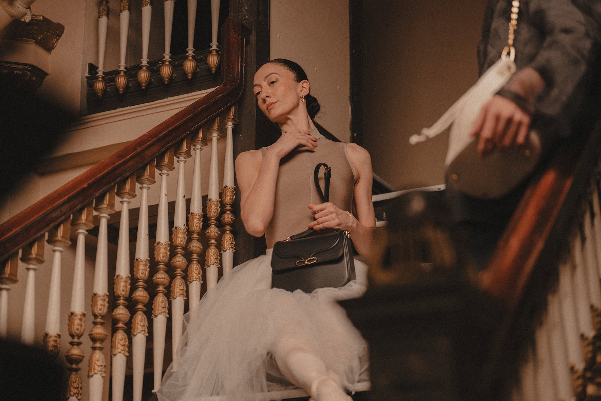 Dancer in a beige top and white tutu holding a black handbag seated on a wooden staircase with white spindles.