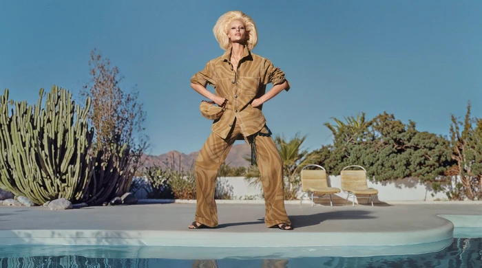 Model with voluminous blonde hair wearing a patterned tan matching set beside a pool and cacti.