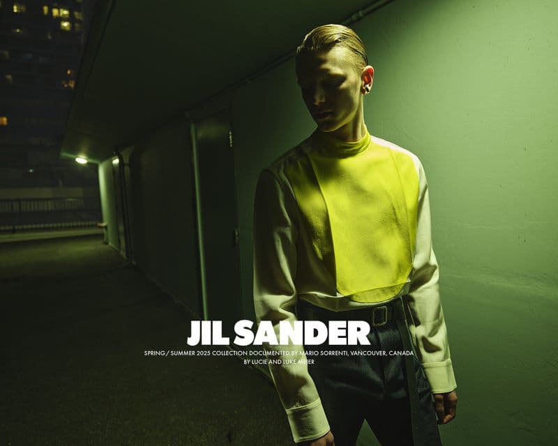 Model in a high-neck yellow and white top standing against a green lit outdoor wall at night