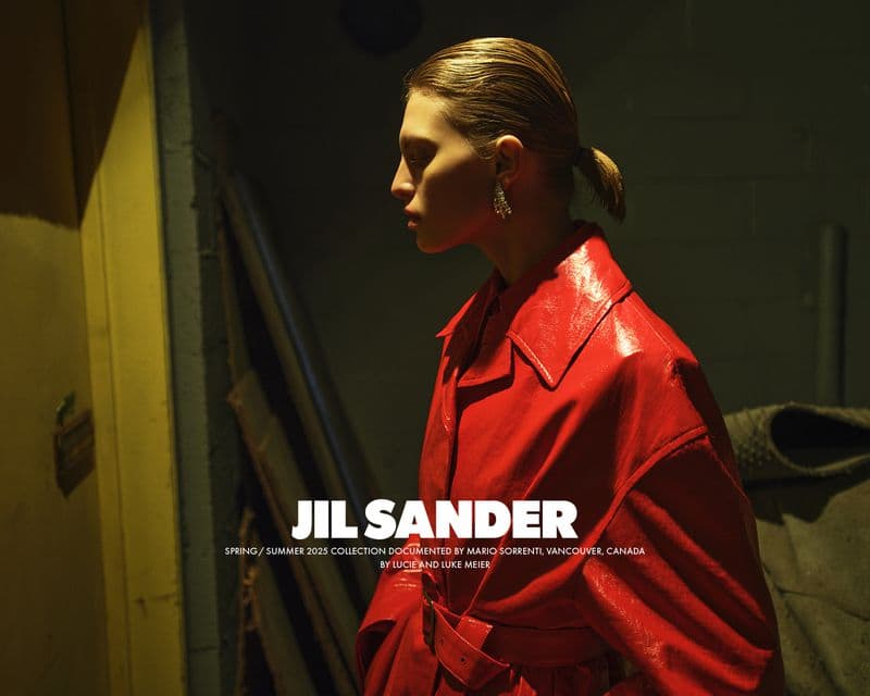 Model with slicked-back hair in a glossy red coat wearing crystal earrings in a dimly lit interior