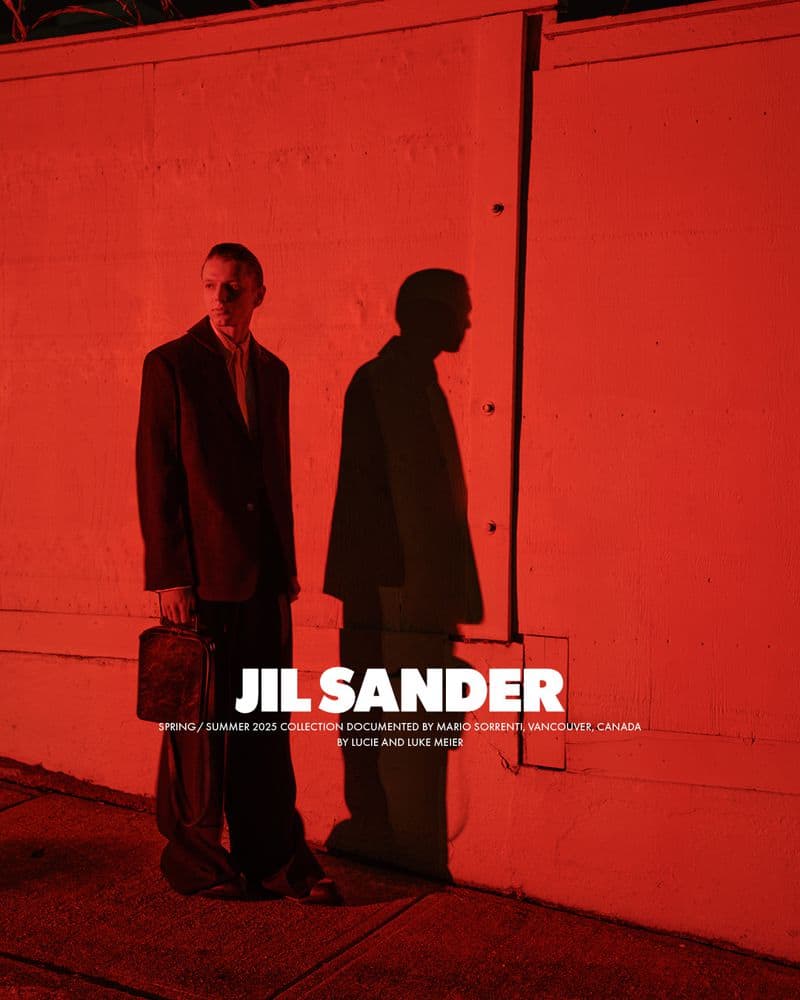 Man wearing a dark jacket and trousers holding a handbag against a red wall with a prominent shadow.