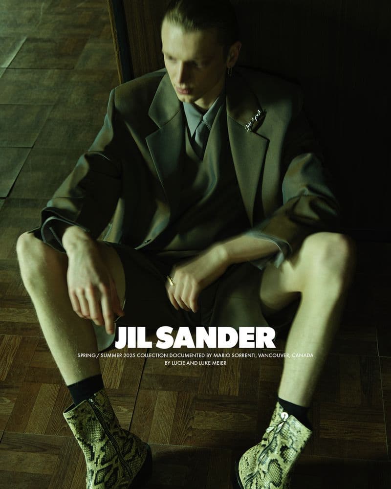 A seated male model wearing an olive green suit jacket, shorts, and patterned boots in a dimly lit interior setting.
