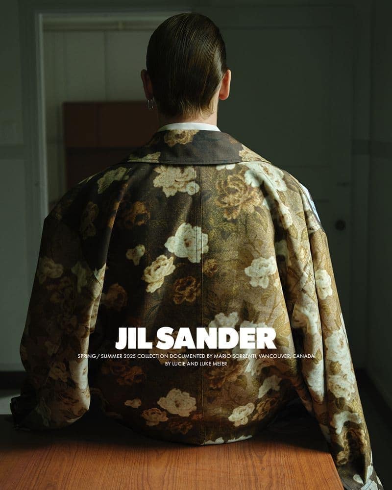 Model seen from behind wearing an oversized floral patterned jacket seated at a wooden table indoors.