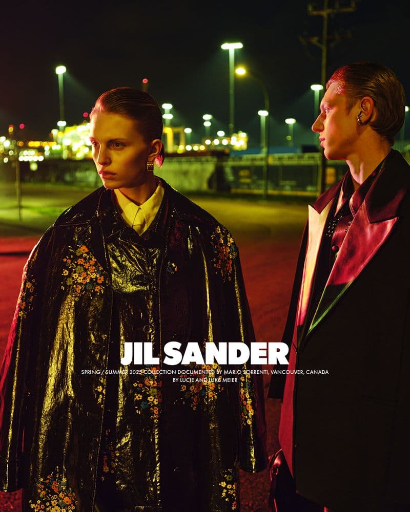 Two models standing outdoors at night under streetlights wearing dark coats for the Jil Sander Spring 2025 ad campaign.