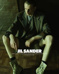 A seated male model wearing an olive green suit jacket, shorts, and patterned boots in a dimly lit interior setting.
