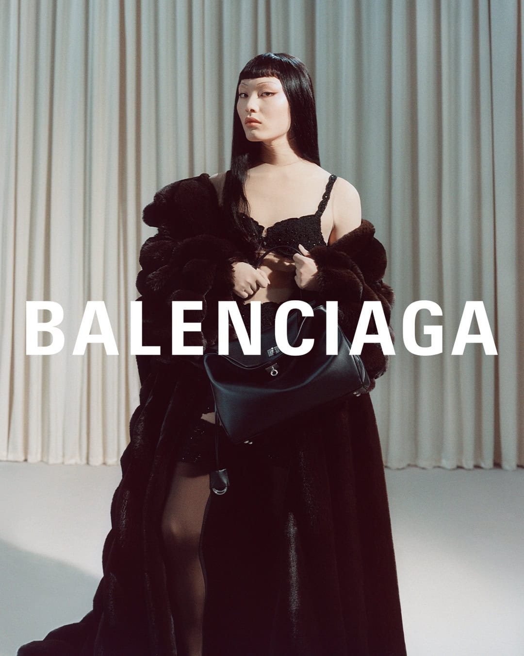 Model in a dark fur coat and black bra holding a leather handbag against a draped curtain background with Balenciaga text