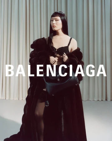 Model in a dark fur coat and black bra holding a leather handbag against a draped curtain background with Balenciaga text