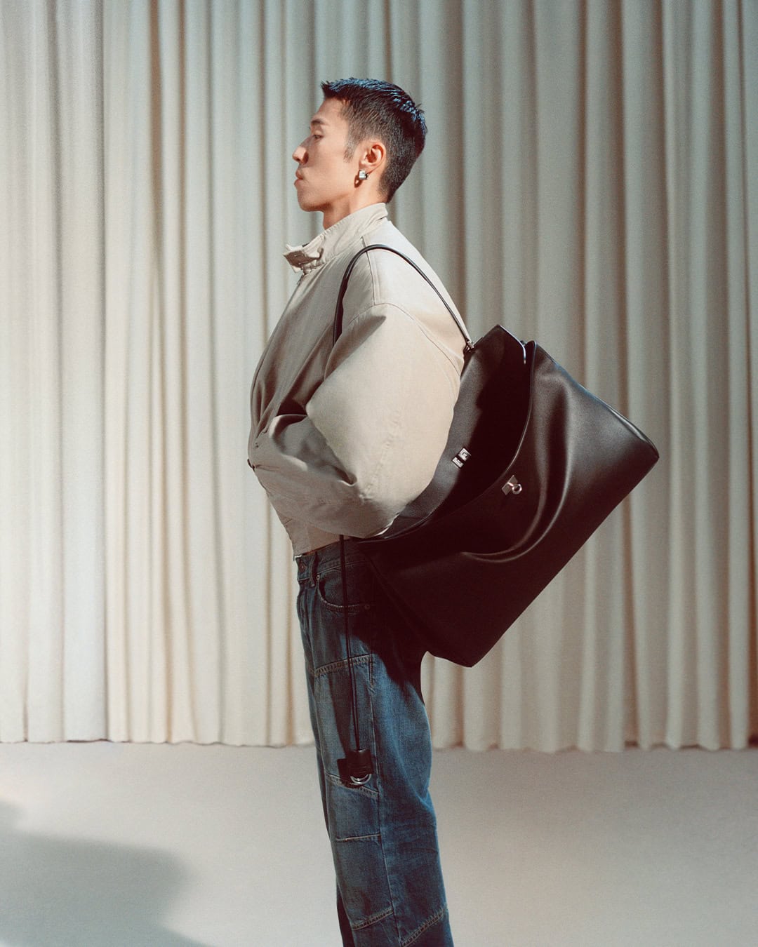 A man wearing a light beige jacket and blue denim jeans standing in profile against a draped curtain background, carrying a large black shoulder bag