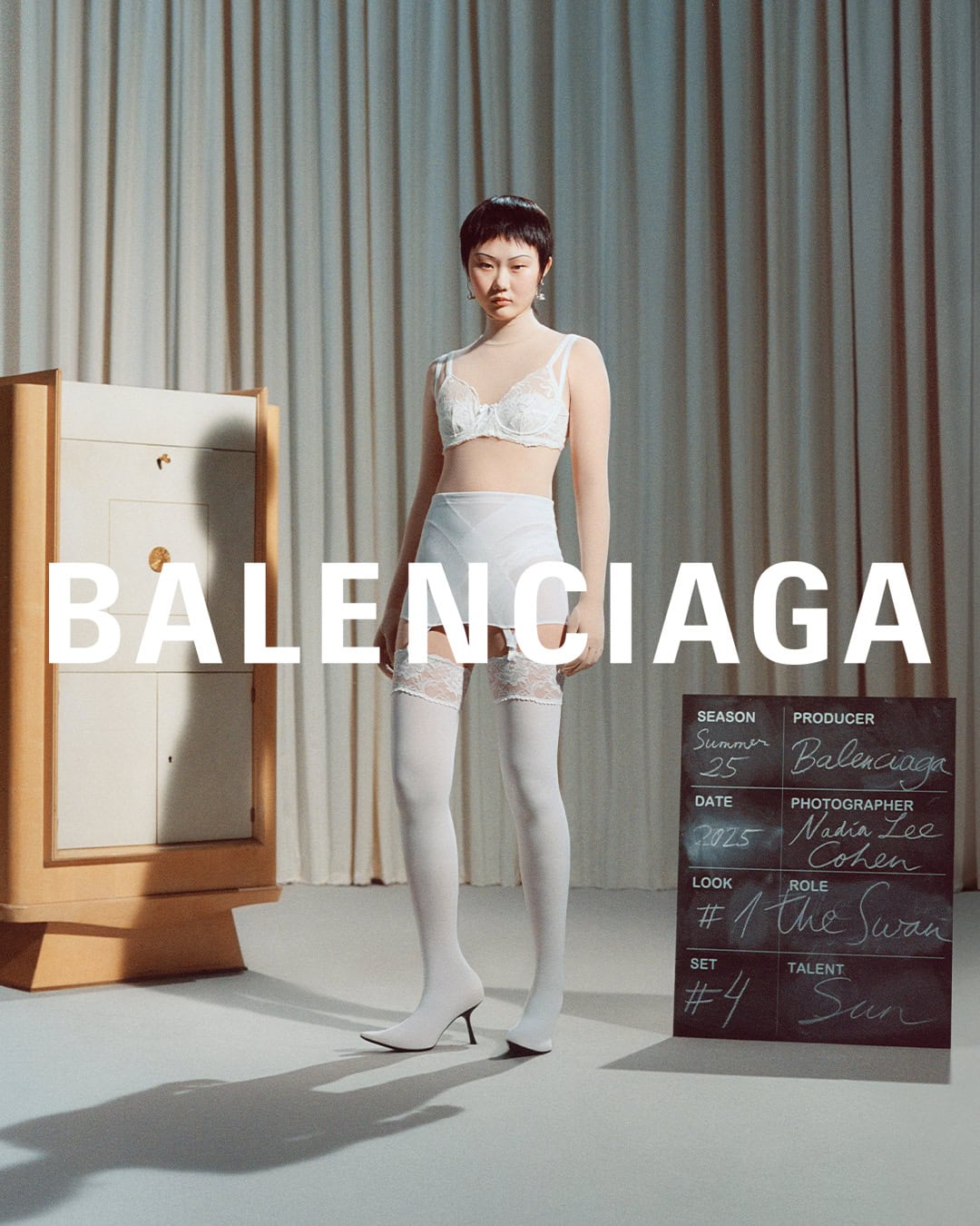 Model wearing white lace lingerie and stockings standing in front of pleated curtains in a studio set by Nadia Lee Cohen for Balenciaga.