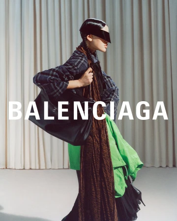Model wearing a plaid shirt and carrying a large black handbag against a pleated curtain backdrop in a Balenciaga campaign.