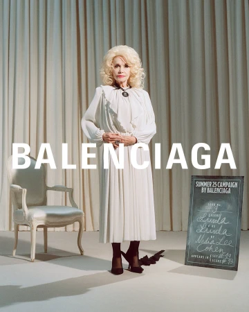 An elderly woman with blonde hair wearing a white pleated dress standing in a room with draped curtains and a vintage chair.