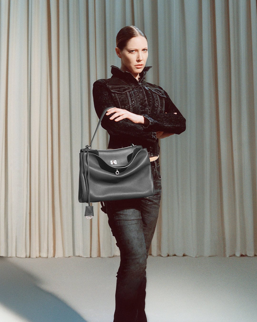 Model standing against vertical curtain background holding a black leather handbag, wearing a dark jacket and dark jeans.