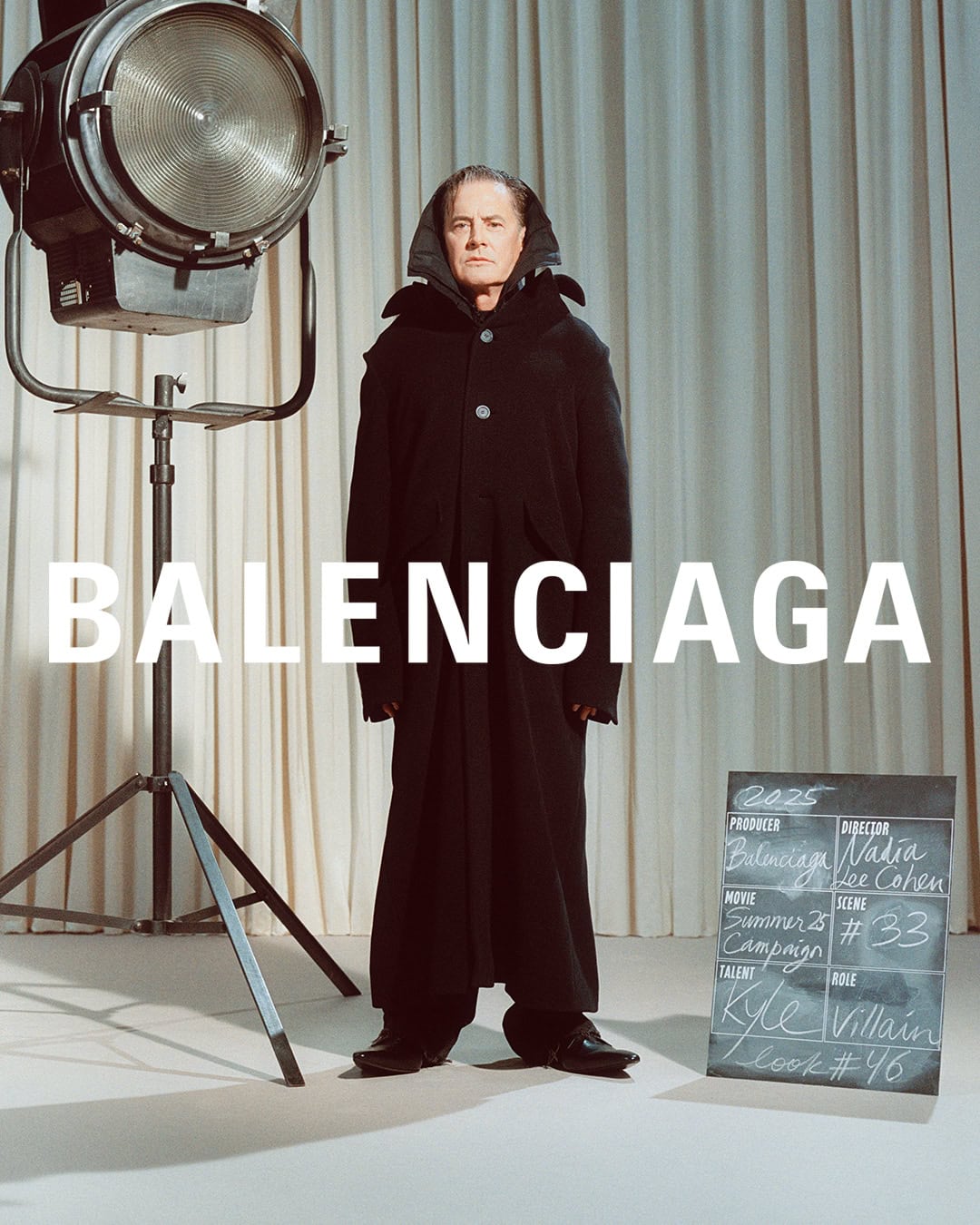 Kyle MacLachlan in a long black coat standing in front of studio curtains next to a studio spotlight and clapperboard