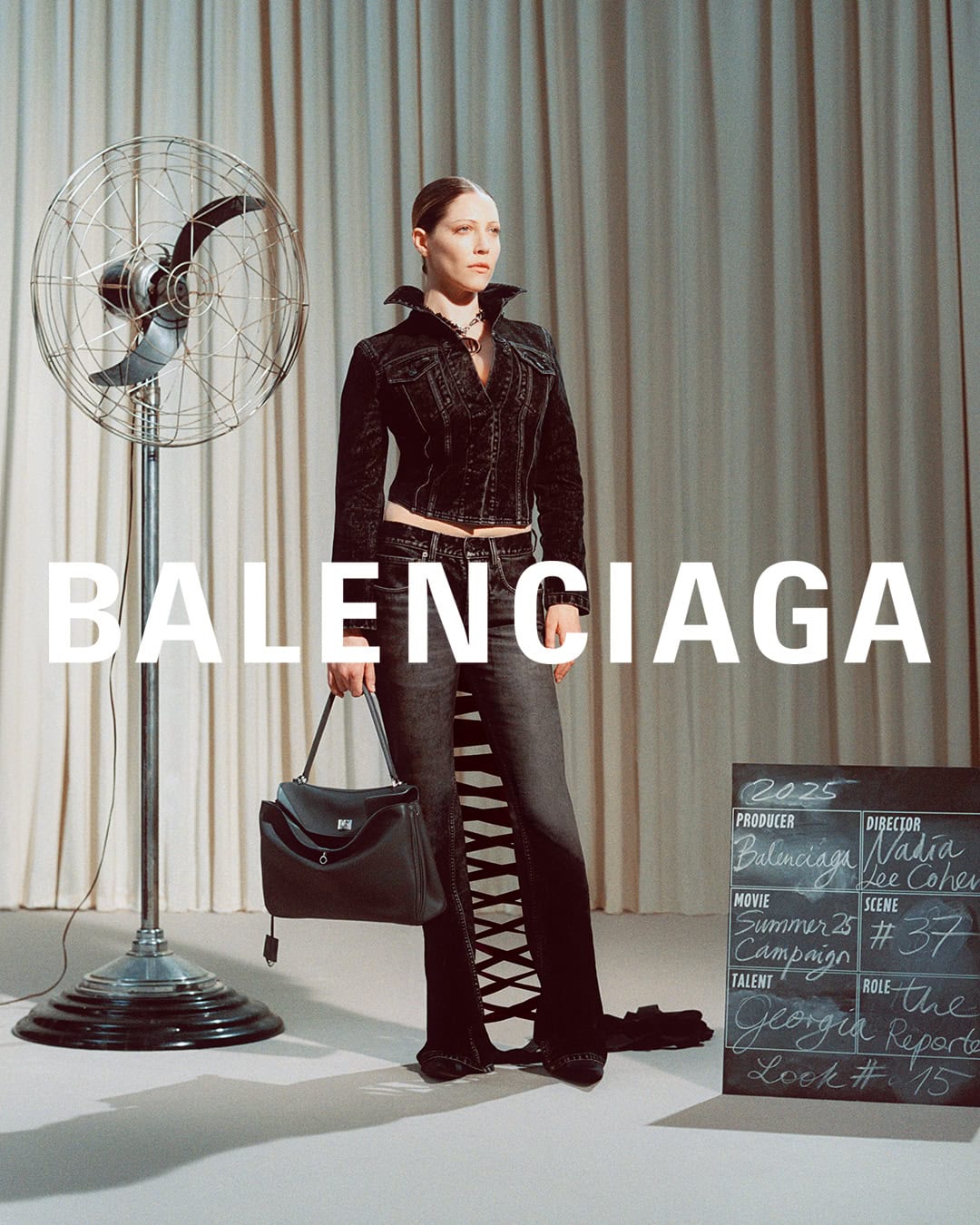 Model standing in dark denim jacket and jeans holding a black handbag next to a vintage fan for Balenciaga