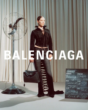 Model standing in dark denim jacket and jeans holding a black handbag next to a vintage fan for Balenciaga
