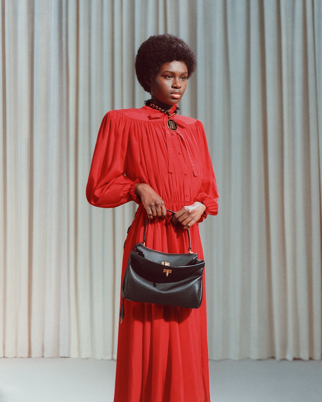 Model in a red dress holding a black handbag standing in front of pleated curtains for Balenciaga