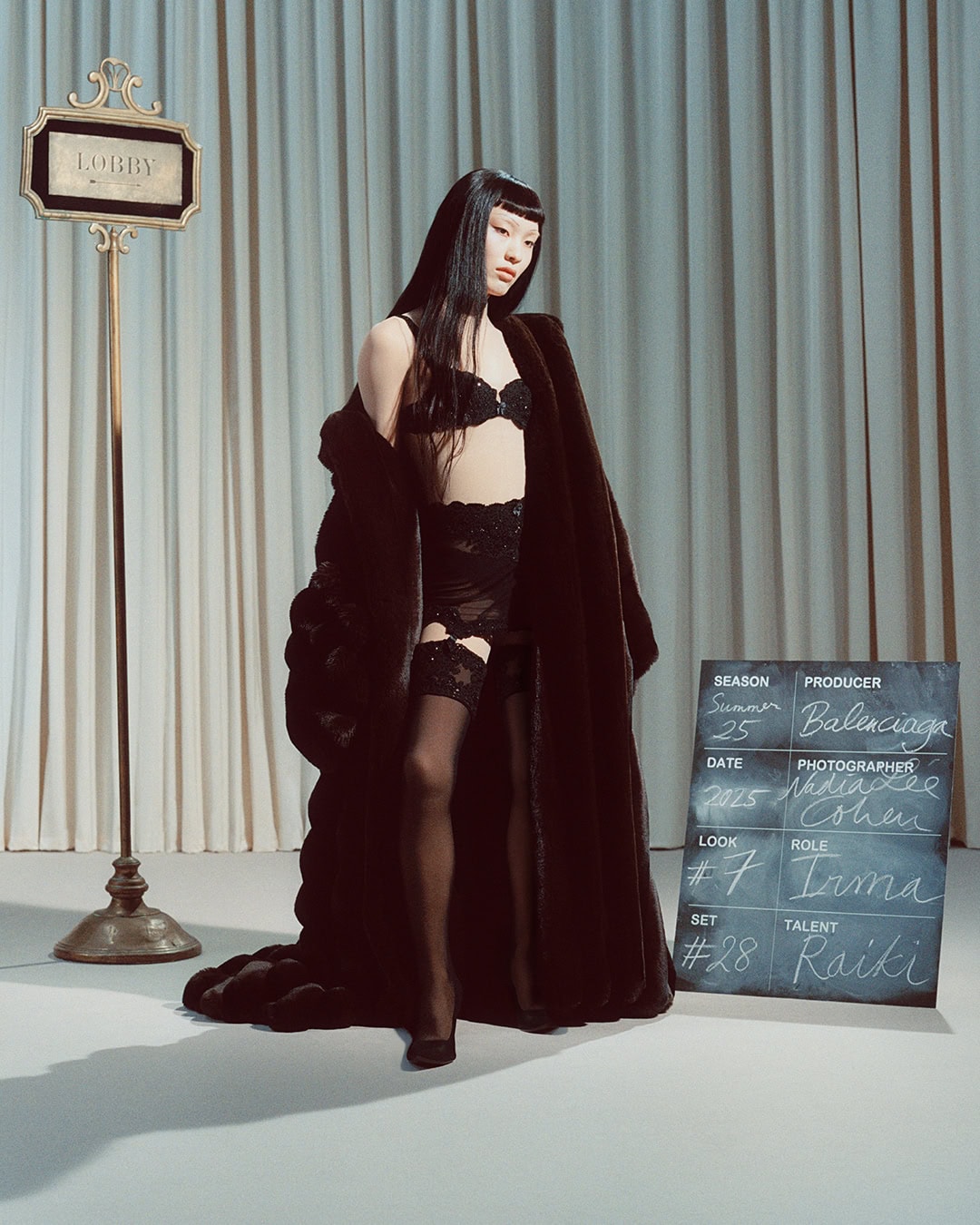 Model in black lingerie and oversized dark coat standing against a draped curtain backdrop with a sign and clapperboard.