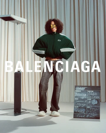 Model wearing a dark green oversized zip jacket and dark jeans standing before draped curtains with a standing light.