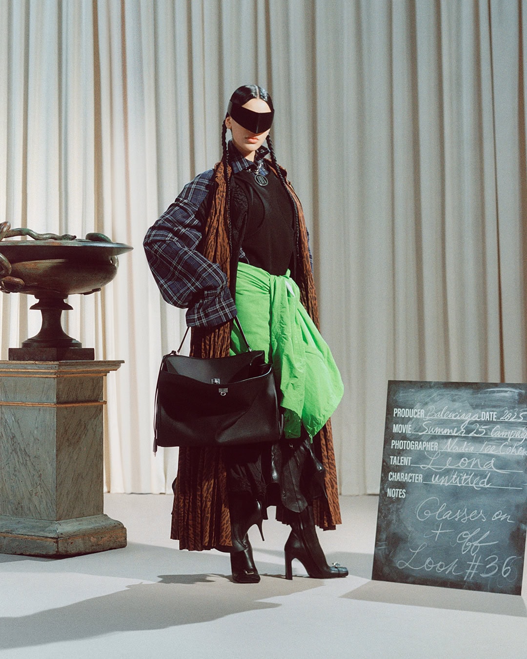 Model in dark wrap-around sunglasses wearing a plaid shirt, brown coat, and bright green wrap holding a black leather handbag.