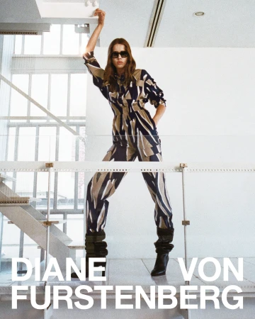 Model wearing a patterned jumpsuit and dark sunglasses standing on a balcony by a glass staircase.
