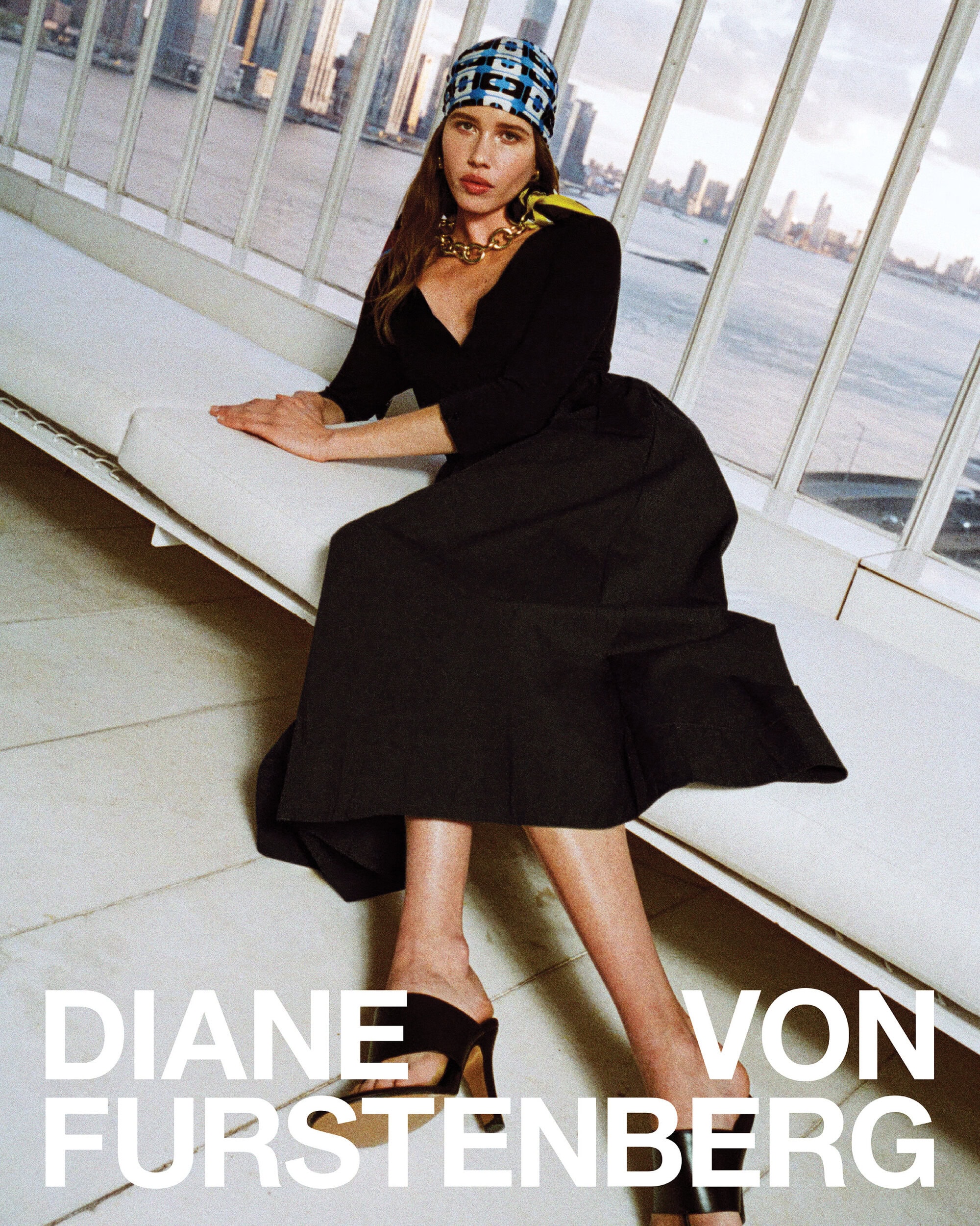 Model seated on a white bench wearing a black dress, patterned headscarf, and gold necklace against a large window.