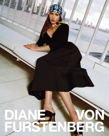 Model seated on a white bench wearing a black dress, patterned headscarf, and gold necklace against a large window.