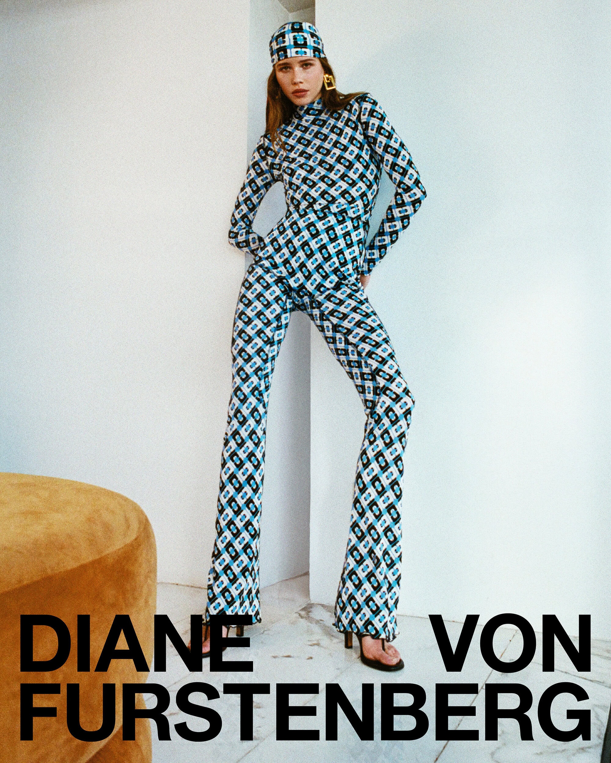 Model wearing patterned blue and black long-sleeve top and trousers with matching headscarf against a white wall.