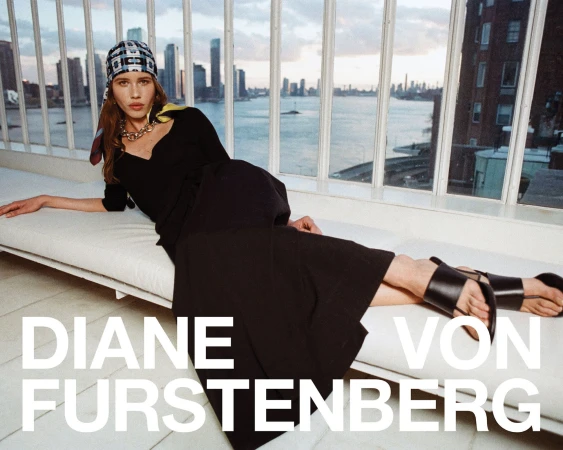 Model in a black dress wearing a patterned headscarf reclining on a white cushioned lounge with a city skyline view behind windows
