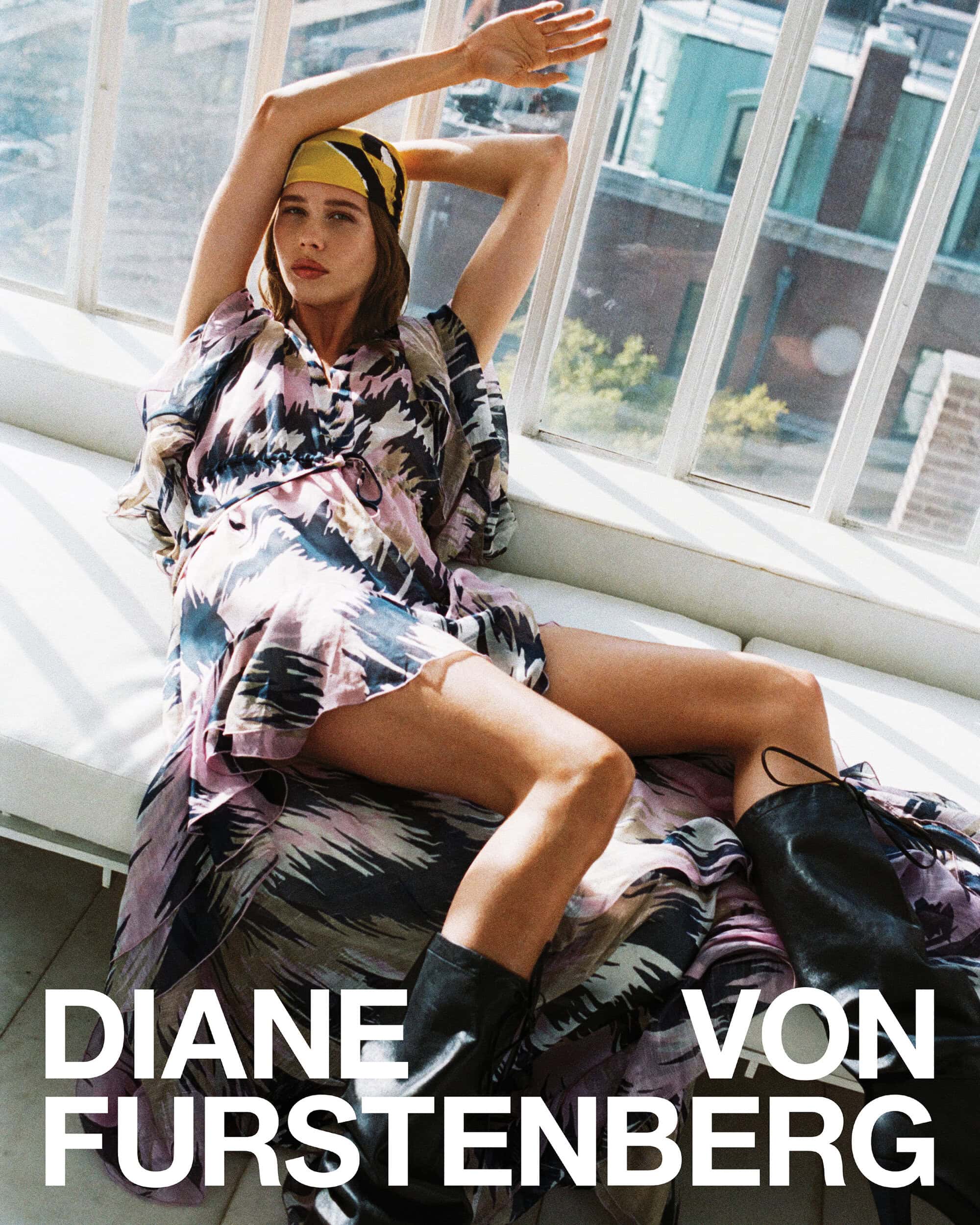 Model reclining in front of large windows wearing a patterned dress, yellow headscarf, and black leather boots.