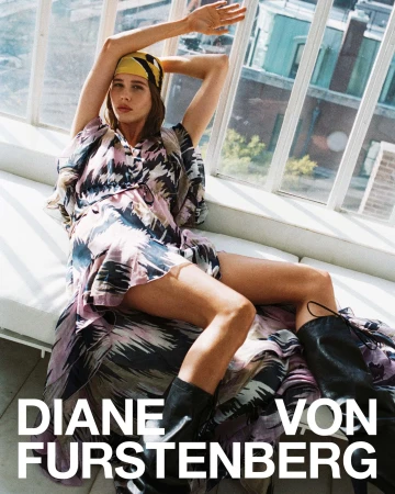 Model reclining in front of large windows wearing a patterned dress, yellow headscarf, and black leather boots.